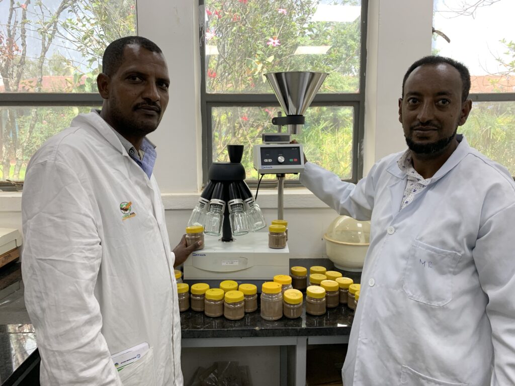 Two men in white lab coats stand in front of an experimental set-up with jars containing various soil samples.